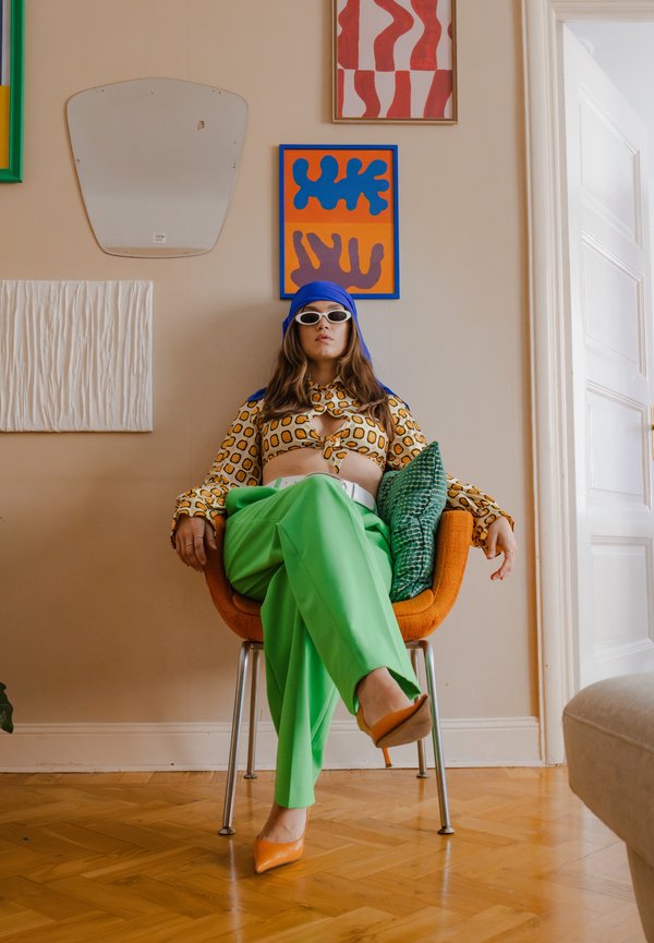 Model zittend op een oranje stoel, gekleed in een gestreepte gele top, groene wijde broek en oranje hakken. Omringd door kleurrijke wandkunst.