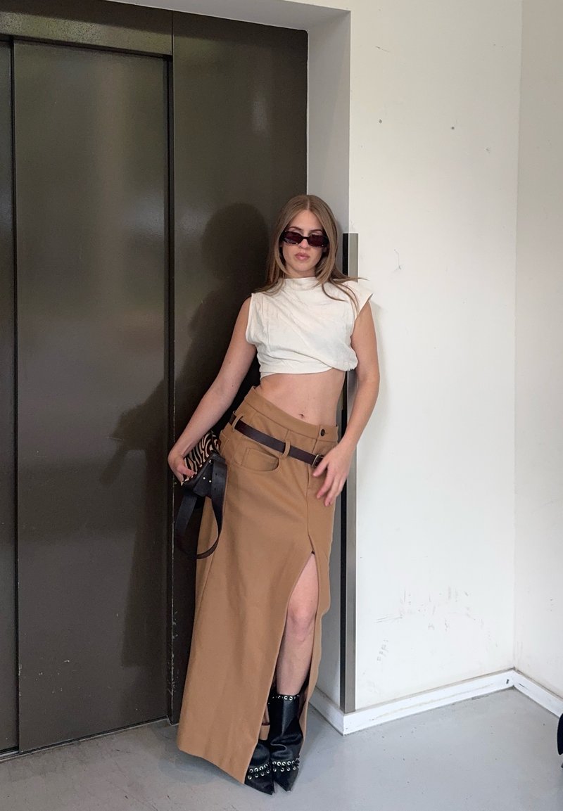 Joven mujer que lleva un top corto sin mangas blanco, falda marrón con una abertura alta, botas negras y gafas de sol, apoyada contra una puerta oscura de ascensor.