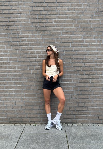 Camisa blanca de botones, pantalones cortos negros, calcetas deportivas blancas y zapatillas plateadas. De pie contra una pared de ladrillos grises, con un pañuelo de cabeza con estampado.