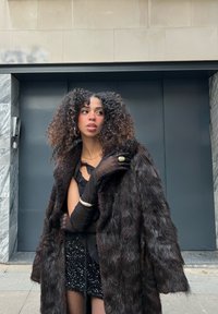 Schwarzer Mantel aus Kunstpelz, schwarze Minirock mit Pailletten und eng anliegende schwarze Bluse, accessoriert mit schwarzen Handschuhen und silbernem Schmuck.