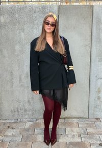 Melns blazera jakas ar garām piedurknēm, caurspīdīga melna svārku ar mežģīņu apakšu, bordo krāsas zeķubikses un tumši sarkanas pulētas augstpapēžu kurpes. Aksesuārs ir zelta aproce.