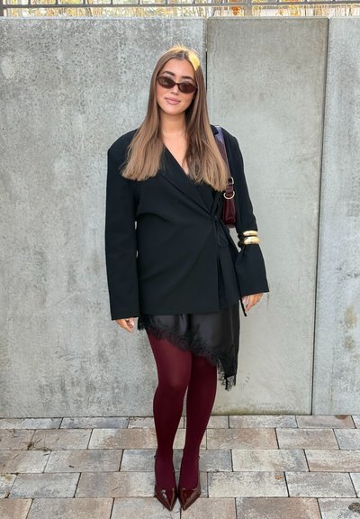 Blazer envolvente negro con mangas largas, falda negra transparente con dobladillo de encaje, medias burdeos y tacones de charol granate. El accesorio es un brazalete dorado.