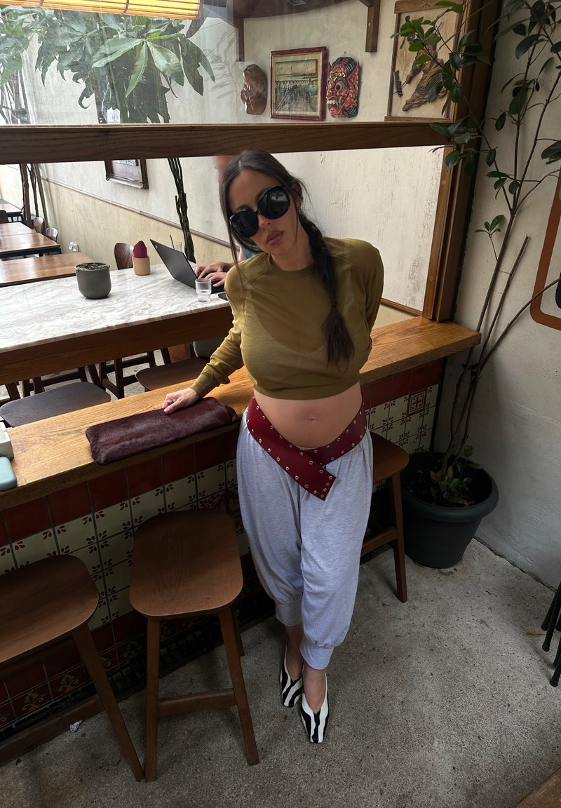 Femme avec une longue tresse foncée et de grandes lunettes de soleil appuyée sur un comptoir en bois dans un café, portant un top court olive, un pantalon gris et des chaussures à motif zèbre.