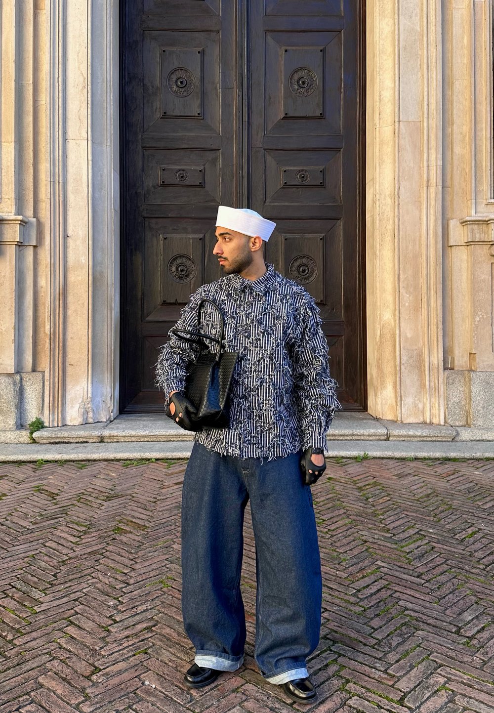 Homme portant un chapeau blanc, une veste à motifs, un jean ample et des gants, tenant un sac noir debout sur un pavement en briques devant une grande porte en bois foncé.