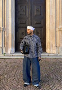 Homme portant un chapeau blanc, une veste à motifs, un jean ample et des gants, tenant un sac noir debout sur un pavement en briques devant une grande porte en bois foncé.