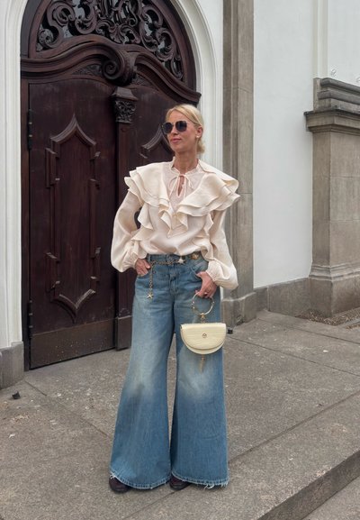 Blusa de crema con volantes y lazo al cuello, jeans de denim desgastado de pierna ancha y un pequeño bolso de marfil con asa circular. Botines marrones. Puertas de madera al fondo.