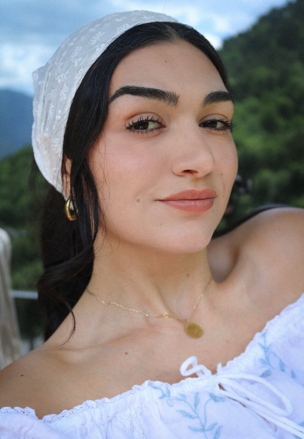 Junge Frau mit dunklem Haar und weißem Kopftuch, trägt goldene Ohrringe und Halskette, im Freien mit Grün und Bergen im Hintergrund.