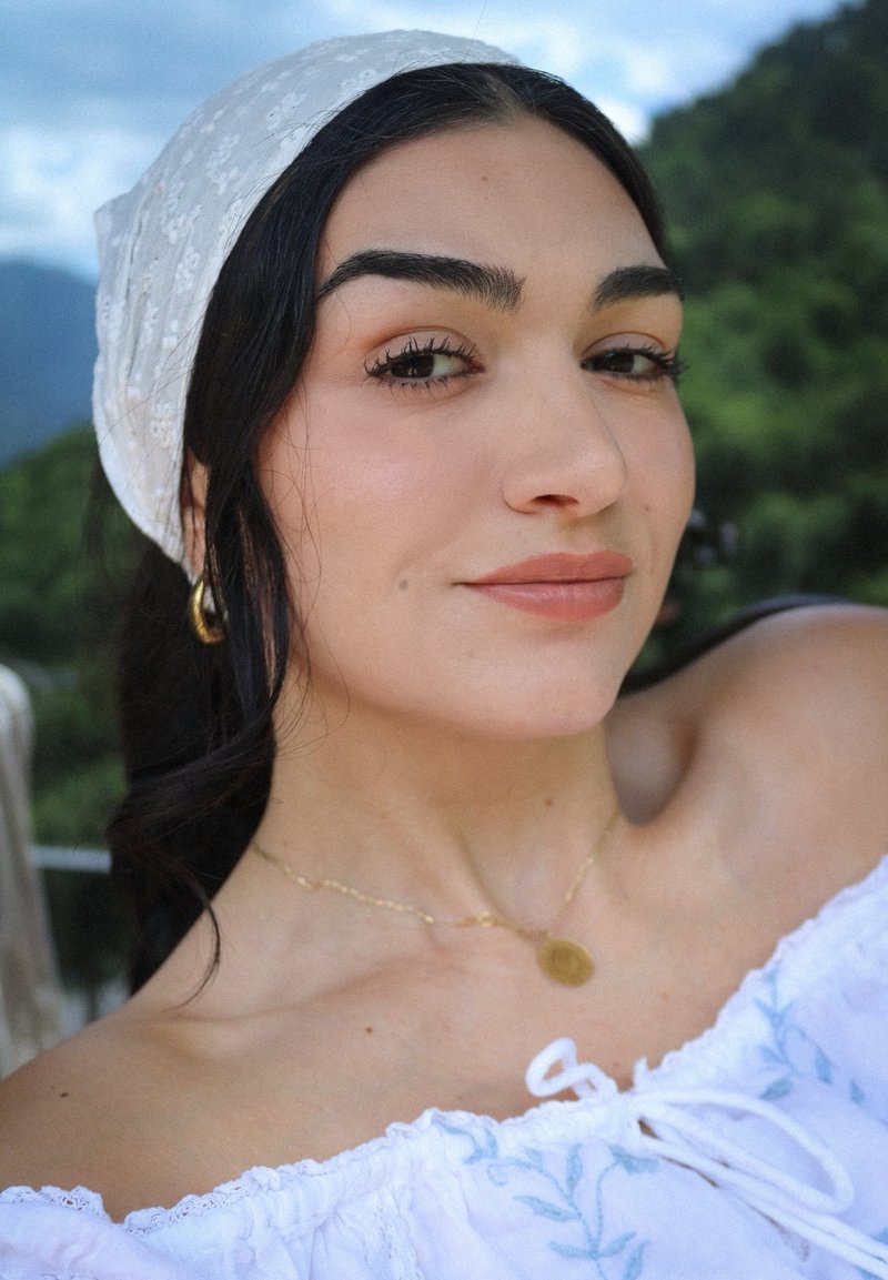 Jeune femme aux cheveux foncés et portant un foulard blanc, avec des boucles d'oreilles et un collier en or, en extérieur avec de la verdure et des montagnes en arrière-plan.