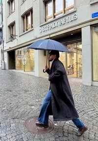 Mees tumeda jope ja teksadega, kes hoiab vihmaumbrella, kõndimas Stradivarius'i poe eest niiskel kivist tänaval.