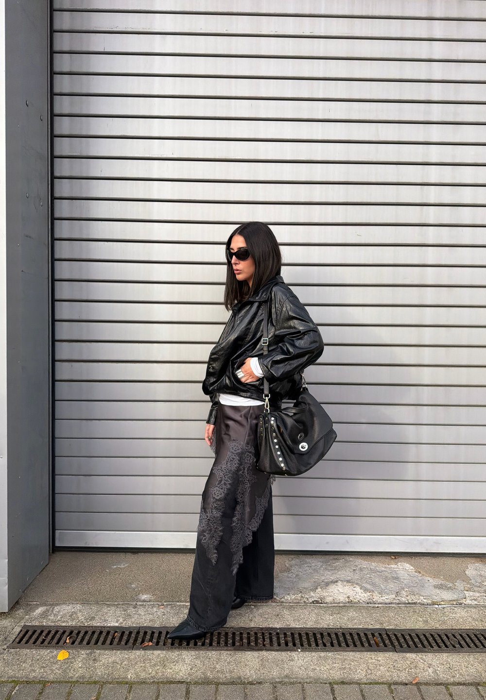 Czarna skórzana kurtka bomberka na białej bluzce, w połączeniu z szerokimi czarnymi spodniami z koronkowym wzorem. Czarna torebka na ramię z ćwiekami.