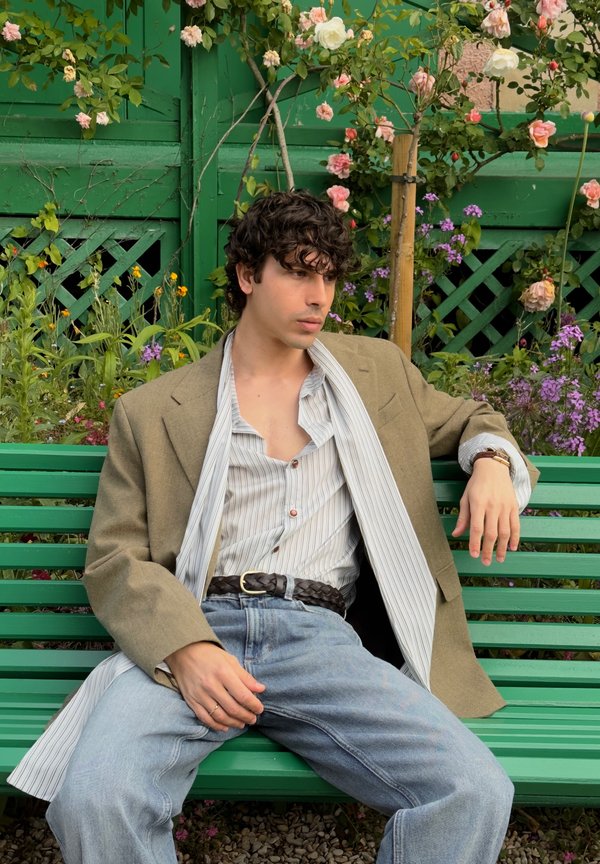 Young man with curly dark hair wearing a beige blazer and striped shirt sits relaxed on a green bench surrounded by blooming flowers.