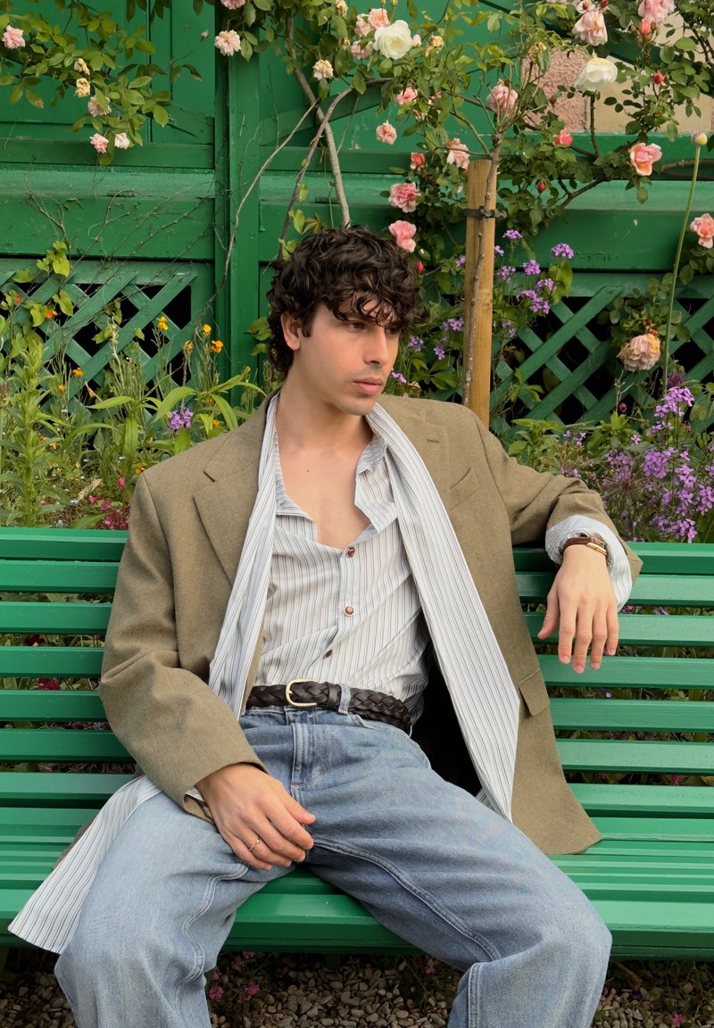 Jeune homme aux cheveux bouclés et foncés portant un blazer beige et une chemise rayée, assis détendu sur un banc vert entouré de fleurs en pleine floraison.