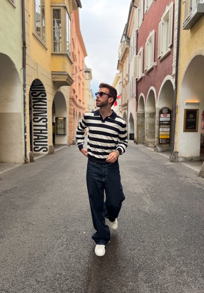 Hombre con gafas de sol y jersey de rayas caminando por una estrecha calle europea con edificios coloridos y arcos a ambos lados.