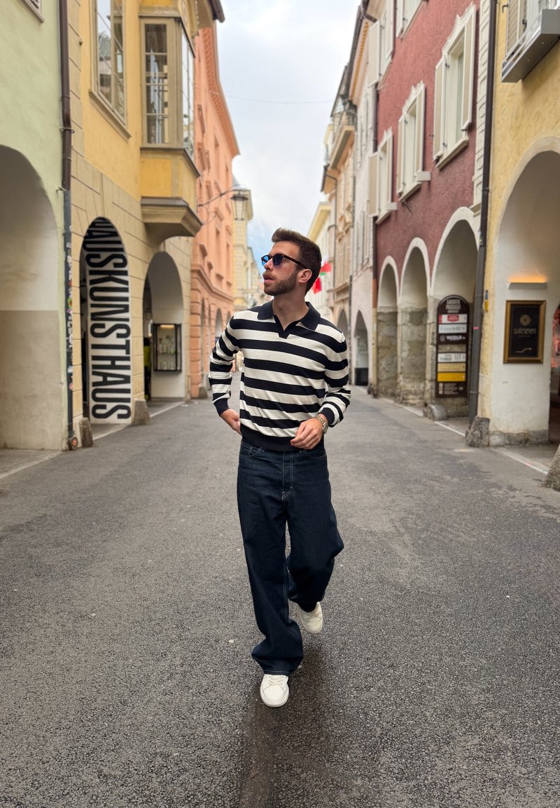 Man wearing sunglasses and striped sweater walking down a narrow European street with colorful buildings and arches on both sides.