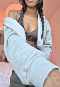 Sweat à capuche gris zippé avec une texture douce par-dessus un soutien-gorge de sport marron. En arrière-plan, une lumière rose douce et une housse de lit blanche froissée.