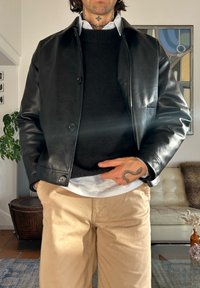Veste en cuir noire avec boutons, portée sur un pull foncé et une chemise blanche. Associée à un pantalon beige clair, présentant une texture lisse.