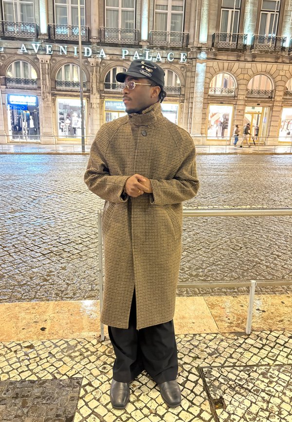 Man in een lange bruine geruite jas, zwarte broek, laarzen, pet en bril staand op betegeld trottoir 's nachts nabij het Avenida Palace-gebouw.