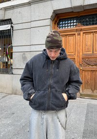 Veste en denim gris foncé avec capuche, poignets élastiques et poches avant. Portée sur un pantalon de jogging gris. Bonnet camo sur la tête.