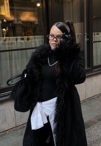 Sort pelsjakke med høj krave, båret over en sort rullekrave. Sort håndtaske med glat tekstur. Langt hår delvist samlet bagud, iført briller.