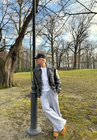 Ung man i svart keps, läderjacka, grå sweatpants och bruna stövlar som lutar sig mot en svart lyktstolpe i en park med lövfria träd.