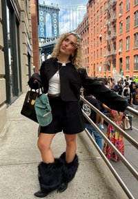 Svart, cropped jakke over en hvit topp, sammen med svarte shorts. Svart, pelsete knelengde støvler, bærende på en svart håndveske med en grønn caps.