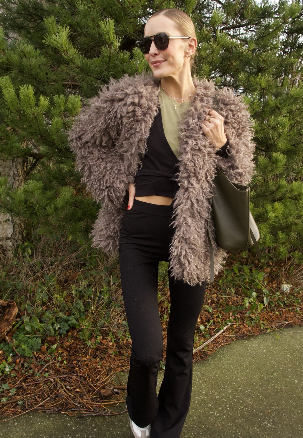 Manteau taupe duveteux, haut noir ajusté, pantalon noir évasé, avec un sac vert. Les accessoires comprennent des lunettes de soleil noires. L'arrière-plan est verdoyant.