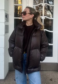Veste rembourrée marron avec col montant, fermeture éclair et poches latérales. Portée sur un pull sombre et un jean bleu clair. Grandes lunettes de soleil.