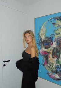 Črna ramenska jakna, siva majica, blond lasje. Ozadje prikazuje barvite umetnine lobanj na modri steni. Vrata z črnim ročajem.