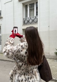 Mākslīgās kažokādas mētelis ar leoparda printu, sarkanie ādas cimdi, brūna rokassoma ar rakstu, gari mati, persona, kas tur viedtālruni pilsētas vidē.