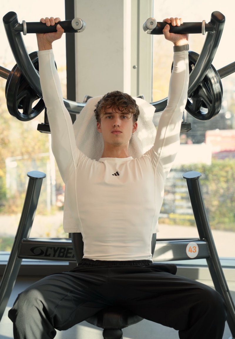 Styrketräningsmaskin med svarta handtag, metallstruktur och en handduk hängd över sätet. Person som lyfter vikter, klädd i en vit tröja med långa ärmar.