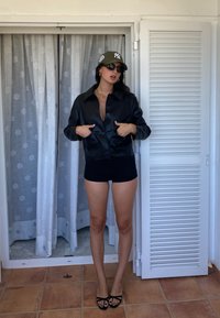 Čierna umelá kožená bunda s gombíkovým zapínaním, nosená cez čierny top a šortky. Zelená čiapka s logom, čierne slnečné okuliare a sandále na opätku.