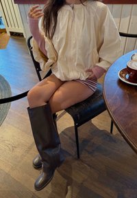 Chaqueta oversize cremosa con textura plisada, puños acanalados rosas, pantalones cortos a rayas y botas de cuero marrón altas, sentado en una mesa de madera.