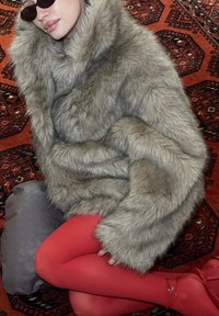 Personne portant un manteau en fourrure gris surdimensionné, des collants rouges, des talons rouges et des lunettes de soleil foncées, assise sur un tapis rouge à motifs avec un sac tressé gris à proximité.