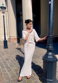 Tenue légère de couleur crème avec un motif texturé ; comprend une blouse à col et une longue jupe ajustée. Des sandales noires complètent le look.