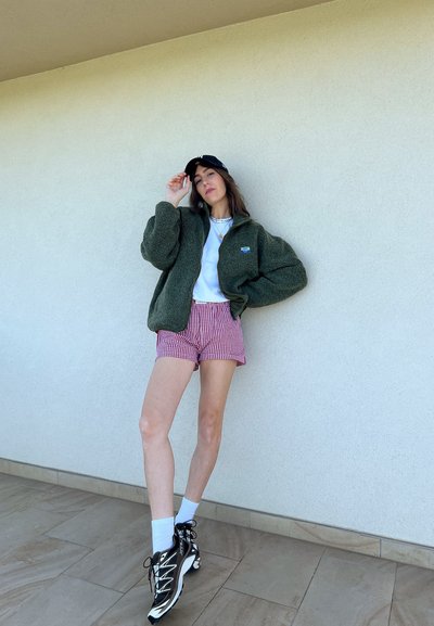 Chaqueta de forro polar verde, camiseta blanca, pantalones cortos a cuadros rojos y blancos, calcetines blancos, zapatillas negras con acentos blancos, de pie sobre una superficie de baldosas.