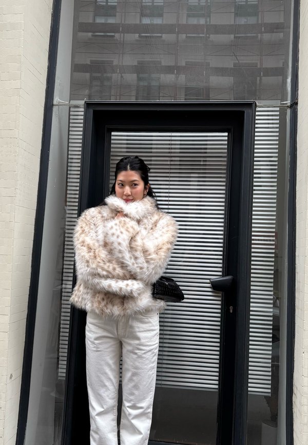 Femme en pantalon blanc et veste en fausse fourrure beige tachetée, debout devant une porte vitrée à cadre noir avec des stores, tenant un petit sac noir.