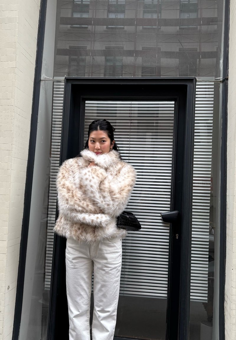 Femme en pantalon blanc et veste en fausse fourrure beige tachetée, debout devant une porte vitrée à cadre noir avec des stores, tenant un petit sac noir.