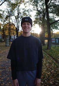 Czarna, krótka bluzka na krótki rękaw noszona na czarnej koszuli z długim rękawem, w połączeniu z czarną dzianą czapką. W tle drzewa i opadłe liście.