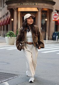 Veste en daim marron sur un haut blanc, pantalon beige taille haute, et baskets blanches. Une casquette beige et une écharpe à motifs ajoutent des détails.