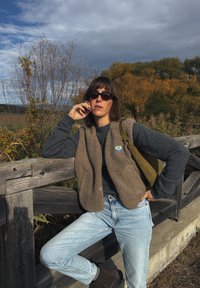 Colete em lã castanho, suéter cinzento, jeans azuis claros e botas castanhas escuras. Saco verde pendurado ao ombro. Corrimão de madeira e folhagem de outono ao fundo.