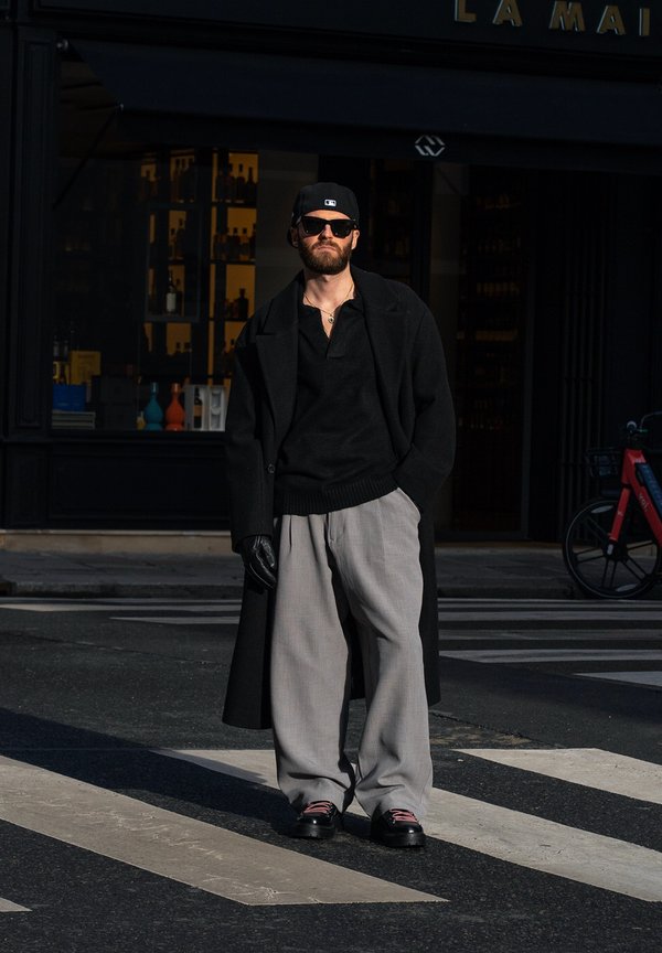 Hombre con abrigo negro, gafas de sol, gorra, guante de cuero y pantalones anchos grises de pie en un paso de peatones frente a una tienda oscura.
