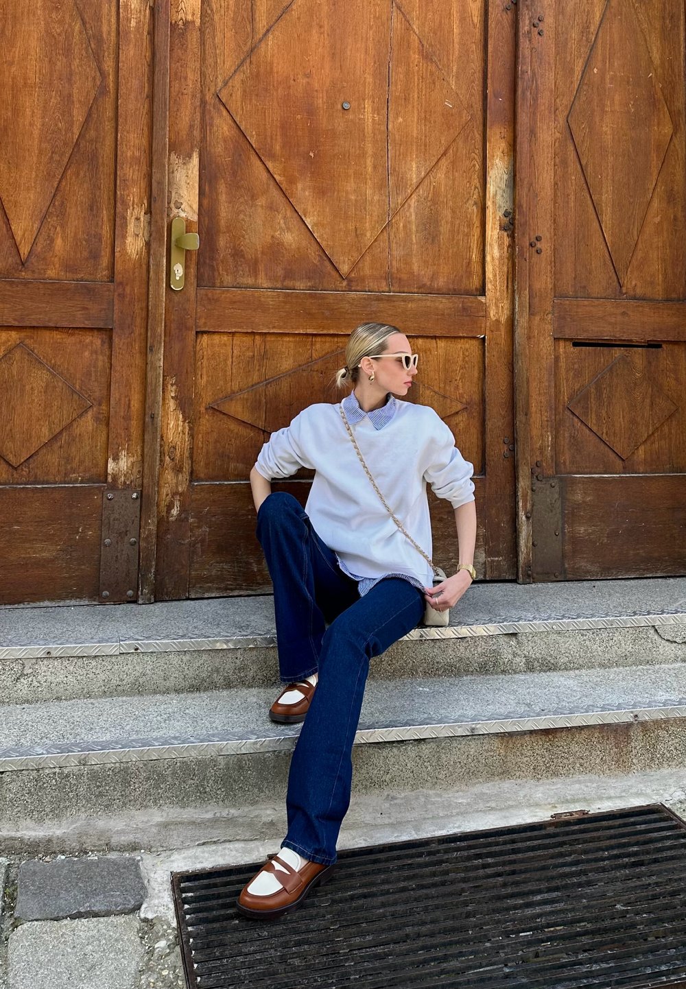 Λευκό oversized φούτερ πάνω από μπλε πουκάμισο με γιακά, μπλε τζιν, καφέ παπούτσια με ύψος και λευκές λεπτομέρειες, καθισμένο σε πέτρινα σκαλοπάτια. Ξύλινη πόρτα στο φόντο.