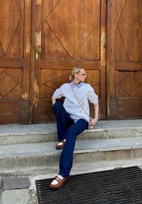 Λευκό oversized φούτερ πάνω από μπλε πουκάμισο με γιακά, μπλε τζιν, καφέ παπούτσια με ύψος και λευκές λεπτομέρειες, καθισμένο σε πέτρινα σκαλοπάτια. Ξύλινη πόρτα στο φόντο.