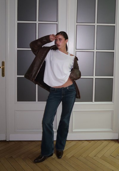 Mujer posando en interiores con una chaqueta de cuero marrón oscuro, top blanco sin hombros, vaqueros oscuros y botas marrones frente a puertas de cristal esmerilado.