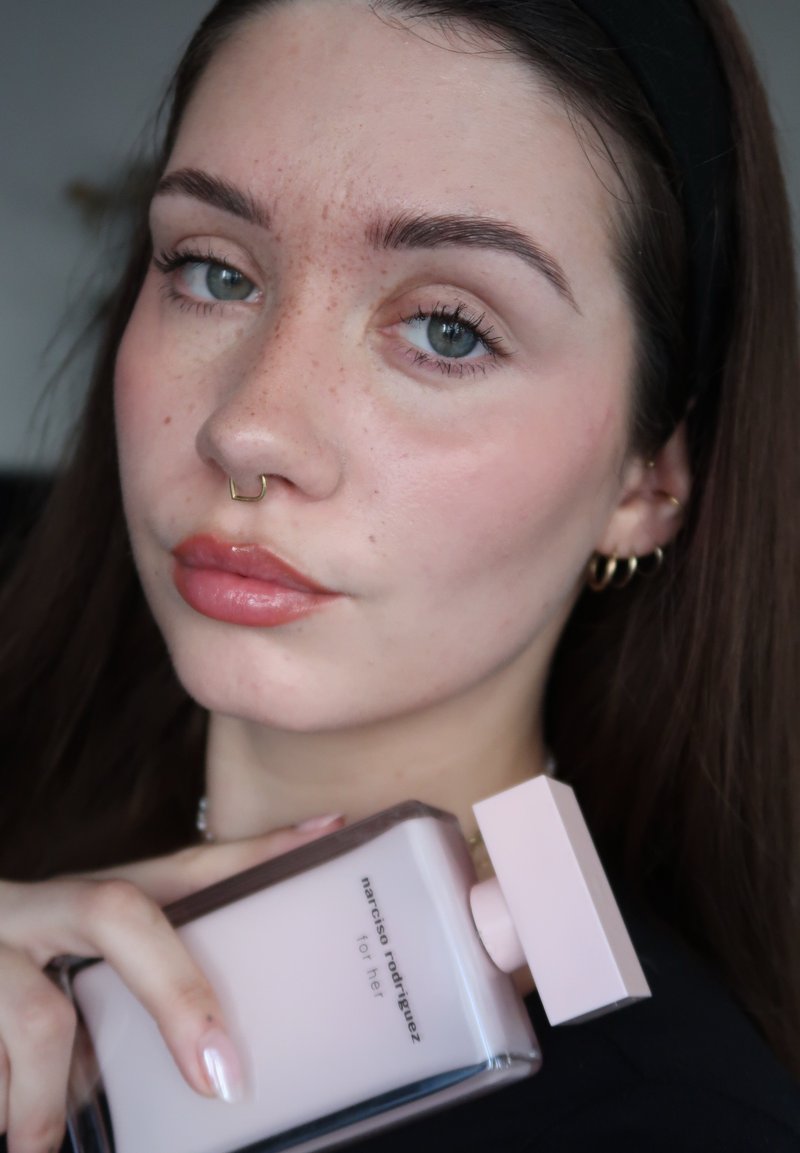Jeune femme avec un anneau de septum et des boucles d'oreilles tenant un flacon rose de parfum Narciso Rodriguez "for her" près de son visage.