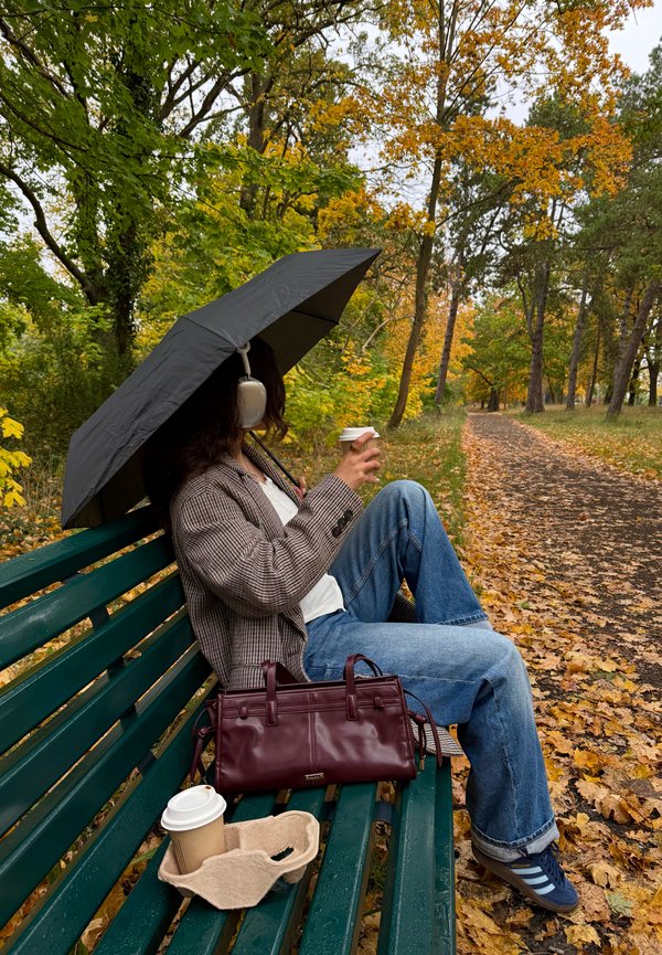 A seated person under a black umbrella is holding a cup of coffee. They are wearing headphones, a checked blazer, and blue jeans, with a burgundy handbag nearby.