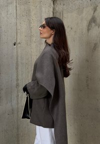 Grå, oversized stofftopp med flytende ermer, kombinert med hvite bukser og svarte hansker, holder en svart håndveske mot en betongbakgrunn.