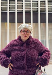 Άτομο που φοράει εκρού μπερέ, κόκκινα γυαλιά και μοβ υφασμάτινο παλτό, στέκεται με τα χέρια στη μέση, κρατώντας μια τσάντα καρό.