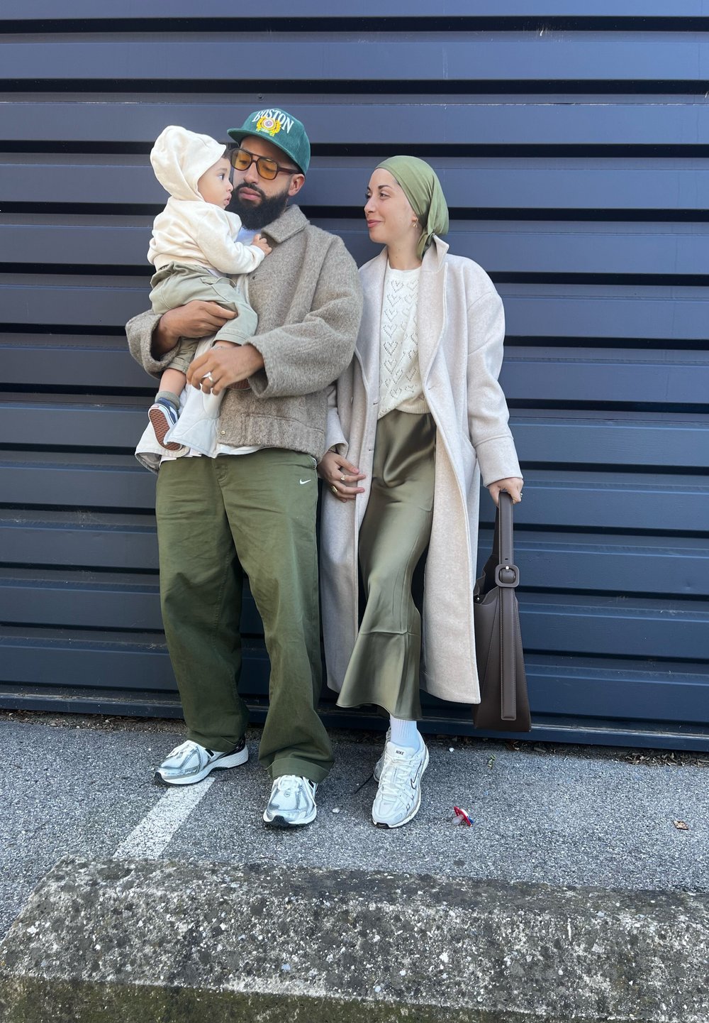 Ενήλικος άνδρας με μπεζ παλτό και γυαλιά κρατάει ένα παιδί με κρέμα ρούχα. Γυναίκα με ανοιχτό παλτό και ελιά χρώμα παντελόνι στέκεται δίπλα, κρατώντας μια καφέ τσάντα.