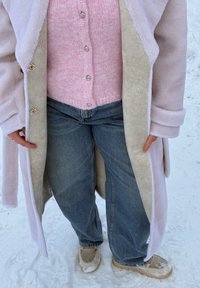Personne portant un manteau duveteux rose clair par-dessus un pull rose boutonné, un jean bleu ample et des chaussures beige, debout sur un sol recouvert de neige.